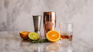 Flat lay of mixology tools and personalized cocktail accessories including shaker, jigger, whiskey glass, and citrus slices on marble background.