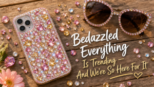 Flat lay on a rustic wood surface — a finished bedazzled phone case, scattered rhinestones in pink, clear, and gold, a pair of bedazzled sunglasses, and a few loose gems catching the light. Warm natural daylight, bright and summery. Text overlay reads: "Bedazzled Everything Is Trending — And We're So Here For It" in a fun, casual font in white or soft gold.